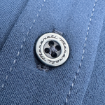 Close-up of a metallic button engraved with the Maserati name, stitched onto blue fabric with visible seams.