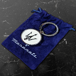 Silver keychain with Maserati trident logo on a white circle, placed on a blue velvet pouch with Maserati name embroidered.